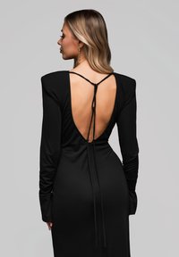 Abito lungo nero con maniche, scollatura profonda sulla schiena, chiusura con lacci regolabili e texture liscia. Design semplice con spacchi laterali.