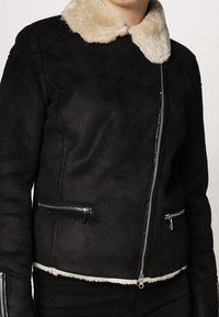 Svart shearlingskinnjacka med en fuskpälskrage, med silverdragkedjor på fickorna och framtill, och en slät textur med en figursydd design.