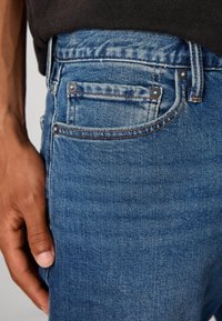 Vaqueros de mezclilla azul con un corte clásico, que cuentan con un diseño de cinco bolsillos, costuras en color cobre y detalles de botones plateados en la cinturilla y los bolsillos.