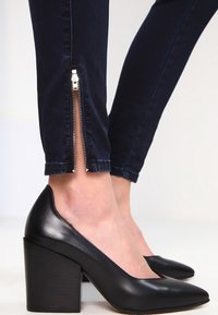 Black leather high-heeled shoes with a pointed toe, paired with dark blue jeans featuring a silver zipper at the ankle.