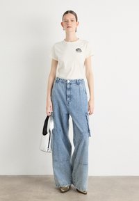 Beige cotton t-shirt with a small black logo, paired with light blue oversized denim cargo pants. Carrying a silver handbag, wearing gold shoes.
