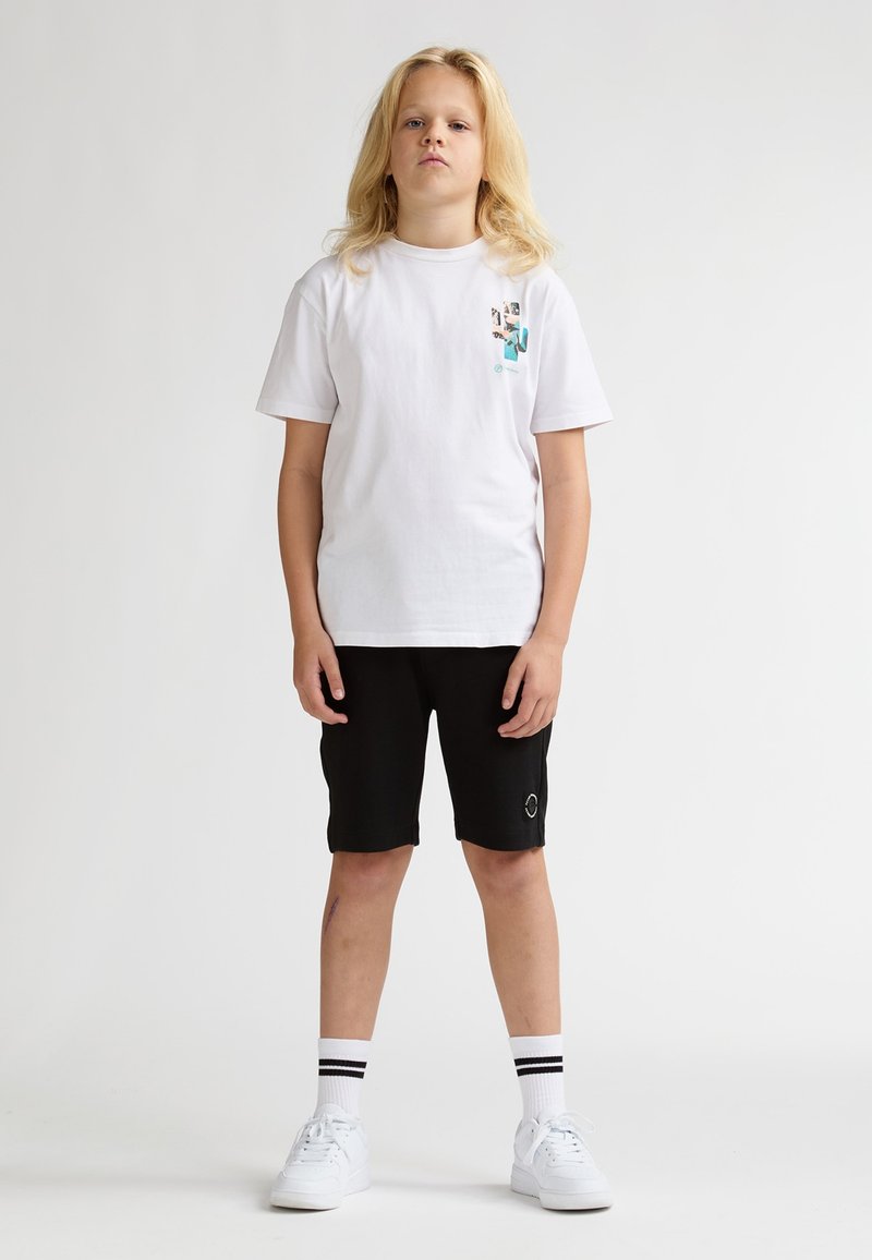 Camiseta blanca de algodón con un diseño gráfico en el pecho, combinada con pantalones cortos negros, zapatillas blancas y calcetines a rayas. Look sencillo y casual.