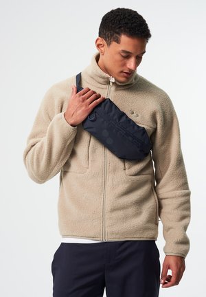 Navy fabric sling bag with a smooth texture, featuring a zipper closure and adjustable strap, worn across the chest over a beige fleece jacket.