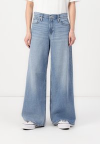 Jeans in denim azzurro chiaro a gamba larga con vita alta, dotati di cinque tasche e texture leggermente sbiadita, abbinati a sneakers bianche.