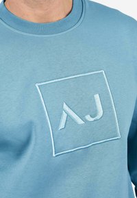 Sudadera azul claro de algodón. Presenta un logo cuadrado bordado "AJ" en un azul ligeramente más oscuro. Cuello redondo y textura suave.