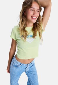 Jasnozielona krótka koszulka z krótkimi rękawami i okrągłym dekoltem, z niebiesko-białym wzorem graficznym. Połączona z jasnoniebieskimi dżinsami.