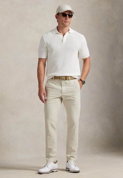 Λευκό πόλο μπλουζάκι με κοντά μανίκια και υφή υφάσματος, μπεζ παντελόνι, λευκά παπούτσια με καφέ λεπτομέρειες, ανοιχτόχρωμο καπέλο και γυαλιά ηλίου.