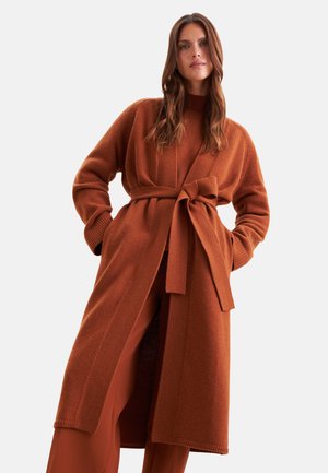 Donna con capelli castani lunghi che indossa un cappotto di lana color ruggine con cintura su pantaloni a gamba larga abbinati, su uno sfondo chiaro e uniforme.