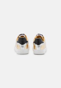 Sneaker con un design metallico oro e argento, con un tallone nero texturizzato, suola bianca e interno giallo.