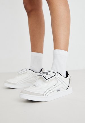 Vita sneakers med svarta accenter och en kardborreband, med en strukturerad ovandel och sidostygn. Bärs med ribbade vita strumpor.