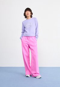 Lawendowa bluza Nike z długim rękawem i wyrazistym logo, zestawiona z jaskrawo różowymi spodniami dresowymi. Szaro-białe sneakersy dopełniają strój.