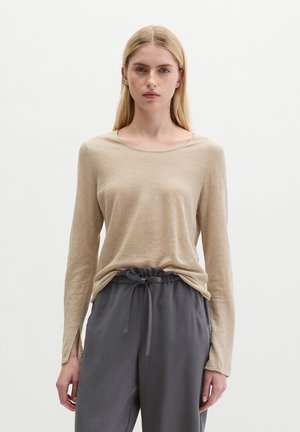 Kobieta o prostych blond włosach, ubrana w beżowy top z długim rękawem z rozcięciami przy nadgarstkach oraz luźne szare spodnie z troczkiem, stojąca na białym tle.