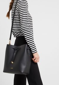 Sac seau en cuir noir avec une fermeture à cordon, une seule bandoulière et des accents en métal doré. Texture lisse, design minimaliste.