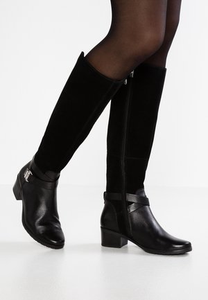 Jambes en collants noirs transparents portant des bottes noires montantes avec talons blocs et brides à boucle, photographiées sur fond blanc.