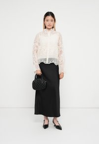 Blusa de encaje con mangas largas, cuello alto y dobladillo ondulado; combinada con una falda maxi de satén negra y un bolso de mano negro texturizado.