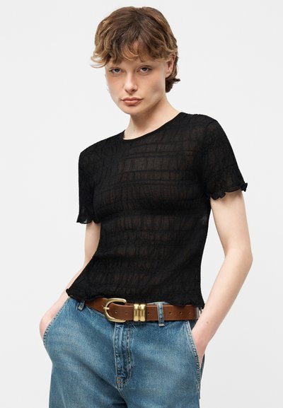 Νεαρό άτομο με κοντά μαλλιά φορούσε ένα διαφανές μαύρο υφασμάτινο κοντομάνικο τοπ, μπλε τζιν και καφέ ζώνη με χρυσή αγκράφα, με τα χέρια στις τσέπες.