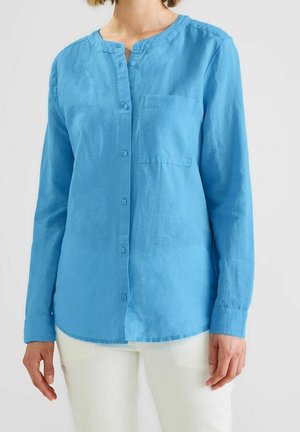 Kobieta ubrana w jasnoniebieską, długą koszulę na guziki z kieszenią na piersi oraz białe spodnie, stojąca na jednolitym tle.