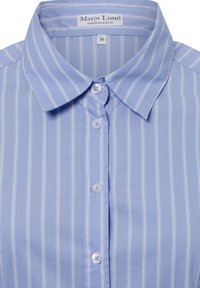 Light blue collared shirt with thin white vertical stripes, featuring a button closure and a label reading "Marie Lund, Copenhagen."