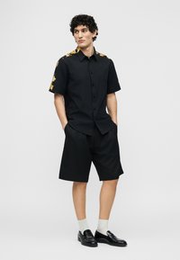 Νεαρός άνδρας με κατσαρά μαλλιά φοράει μαύρο πουκάμισο με κοντά μανίκια και χρυσό μοτίβο, μαύρο σορτς, λευκές κάλτσες και μαύρα μοκασίνια.