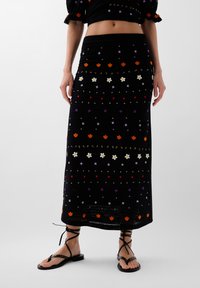 Jupe maxi en tricot noir ornée de broderies florales multicolores, de détails complexes et d'un ourlet en forme de vagues. Assortie avec des sandales noires à lanières.