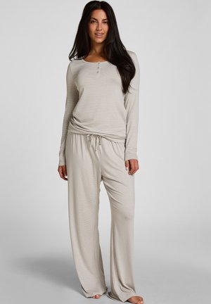 Vrouw met lang donker haar, die een lichtbeige top met lange mouwen en bijpassende brede broek met trekkoord draagt, staat blootsvoets tegen een effen achtergrond.