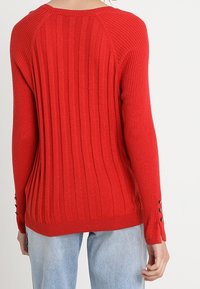Pull rouge côtelé avec manches raglan, présentant des boutons décoratifs sur les poignets et un col rond. Confectionné en maille texturée.