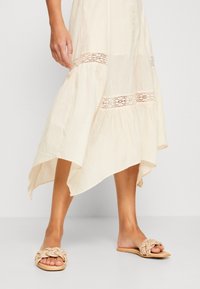 Woman wearing a cream-colored midi skirt with lace inserts and braided beige slip-on sandals, standing on a light gray floor.