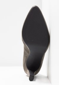Botte à talon en daim gris à bout pointu avec semelle noire texturée, posée à la verticale contre un mur d'angle blanc et un sol blanc.