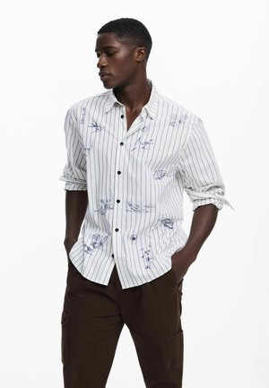 Camisa blanca a rayas con estampado floral azul, cuello camisero y mangas cortas, combinada con pantalones marrón oscuro.