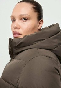 Brązowa ocieplana kurtka z wysokim kołnierzem i kapturem. Posiada zamek błyskawiczny oraz pikowane fragmenty. Gładka, matowa faktura z eleganckim designem.