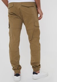 Pantalons cargo kaki avec des poignets élastiques, dotés de plusieurs poches latérales, texture lisse et design ajusté. Portés avec des baskets blanches.
