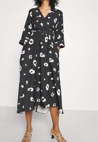 Svart omlottklänning med vitt leopardmönster, V-ringning, trekvartsärmar och midjeband. Flödande tyg med en något asymmetrisk nederkant.