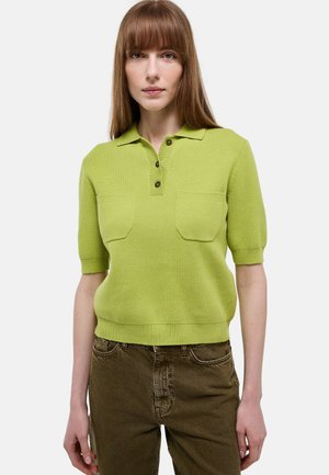 Femme aux cheveux bruns et raides portant un polo en maille vert citron à manches courtes et un pantalon vert olive, debout devant un fond uni.