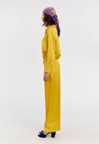 Blouse en satin jaune à manches longues et pantalon large assorti, avec des détails de boutons sur les manches. Associé à des sandales à talons hauts bleu marine et une écharpe à motif.