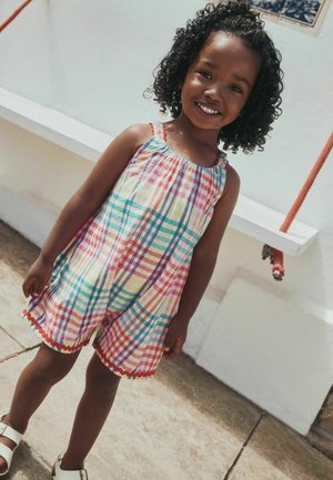 Jeune fille souriante aux cheveux bouclés portant une combinaison sans manches à carreaux colorés et des sandales blanches, debout à l'extérieur sur une surface en béton.