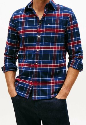 Homme portant une chemise à carreaux bleue et rouge à manches retroussées et boutonnée, les mains dans les poches d’un jean foncé, vu du cou jusqu’aux hanches.