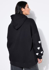 Sudadera negra con una capucha grande, acentos en forma de corazón blancos en las mangas y un corte relajado. Fabricada en material suave.