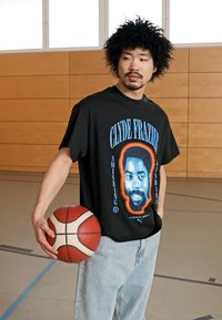 Sort t-shirt med blå og orange grafik af Clyde Frazier's ansigt; parret med lyseblå jeans; holder en basketball.