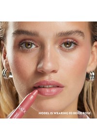 Ceraso per labbra di colore rosa chiaro tenuto vicino alle labbra; il modello presenta una pelle liscia, trucco naturale e orecchini a cerchio argentati. Il testo in basso mostra le informazioni sul prodotto.