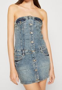 Denim šaty bez ramínek, světlý modrý odstín, přiléhavý střih, dvě náprsní kapsy, knoflíky ve zlatém tónu a detaile šití.