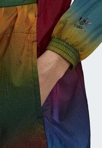 Barevná větrovka se stupňujícími odstíny červené, zelené a modré. Nastavitelné manžety, stažený pas a decentní detail loga.