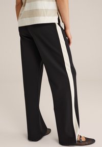Pantalon noir avec une bande latérale blanche contrastante, présentant une coupe décontractée et une texture de tissu lisse. Porté avec des ballerines noires en mesh.