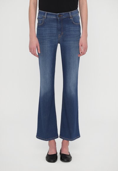 Jean flare en denim de lavage foncé avec une conception à cinq poches, des coutures robustes et une légère décoloration du genou à l'ourlet. Portés avec des ballerines noires.