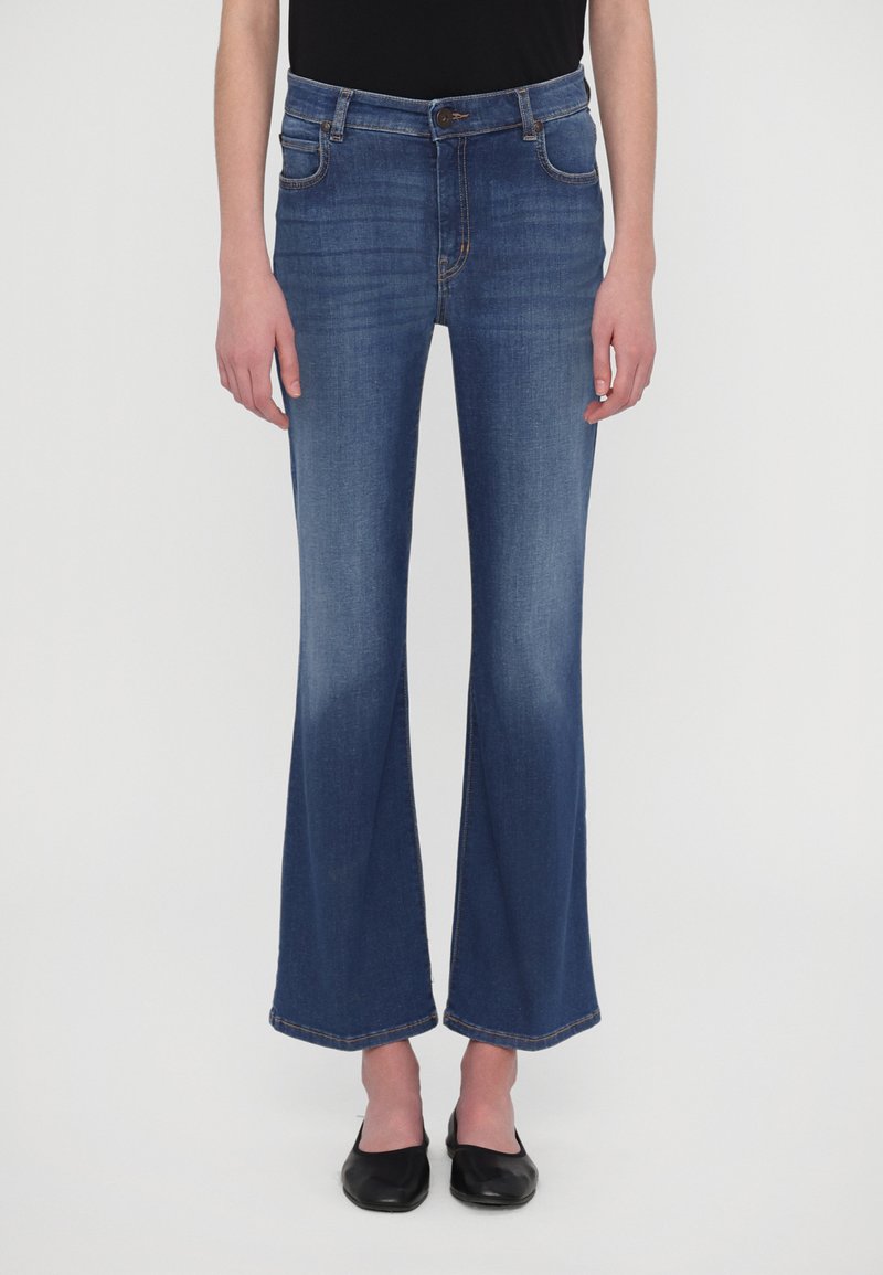 Blugi evazați din denim, de culoare închisă, cu un design cu cinci buzunare, cusături rezistente și o ușoară decolorare de la genunchi până la tiv. Purtați cu pantofi negri.