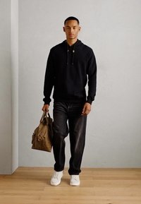 Schwarzer Kapuzenpullover, dunkle Jeans, weiße Sneaker und eine braune Ledertasche. Das Model steht auf einem Holzfußboden vor einem schlichten Hintergrund.