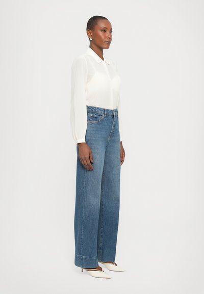 White long-sleeve blouse with collar, paired with wide-leg blue jeans. Model wears white heels, with a smooth texture and classic design.