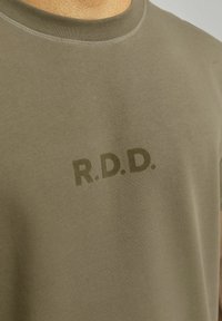 Olivově zelené bavlněné tričko s kulatým výstřihem. Na přední straně má decentní, embosované logo "R.D.D." Hladká textura, ležérní design.