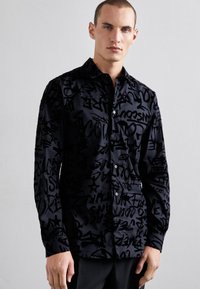 Camicia nera con bottoni, caratterizzata da un motivo testurizzato di diverse lettere e forme in stile graffiti nero, con colletto e maniche lunghe.