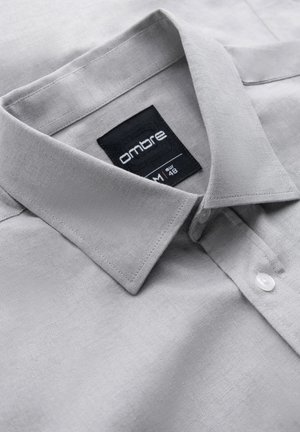 Lichtgrijze button-up overhem met een gestructureerd, visgraatpatroon. Voorzien van een kraag en een label met de tekst "ombre," maat M, EUR 48.