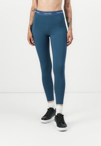 Teal tajice visokog struka s glatkom teksturom, sa pojasom s Calvin Klein logotipom i krojem koji se čvrsto prilagođava tijelu. Uparene s crnim tenisicama.